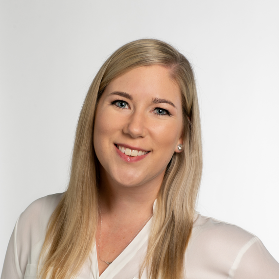 A woman with long blonde hair smiles at the camera, wearing a white blouse and small earrings, in front of a plain white background.