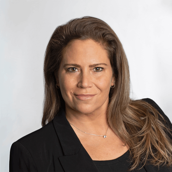 Woman with long brown hair wearing a black blazer and necklace, posing against a plain light background.