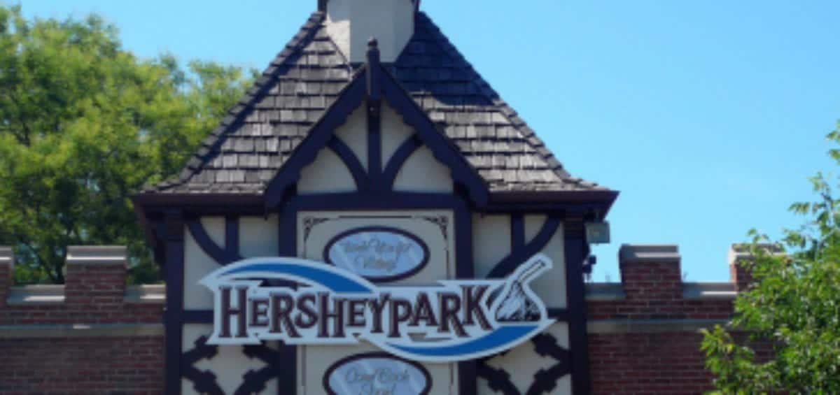 Hersheypark entrance sign featuring a logo with a wave design and a scoop of chocolate. The background shows a building with a small turret and a clear blue sky. An "Exit" sign is below.