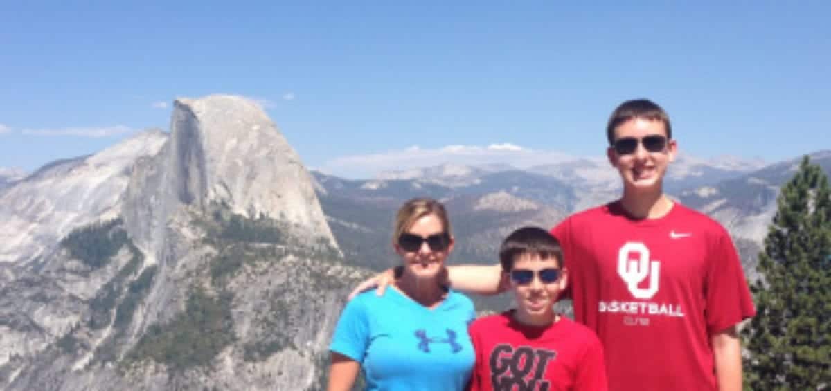 Three people standing together on a mountain overlook with a distinctive rock formation visible in the background. They are dressed in casual clothing and wearing sunglasses.