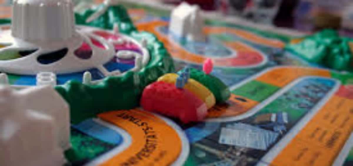 Close-up of a board game showing colorful paths, a spinning wheel, and small car-shaped game pieces. The board is brightly colored with various paths and text.