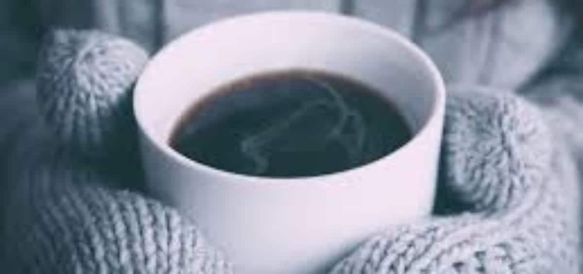 Hands wearing gray knit mittens holding a white mug filled with black coffee, with steam rising from the cup.