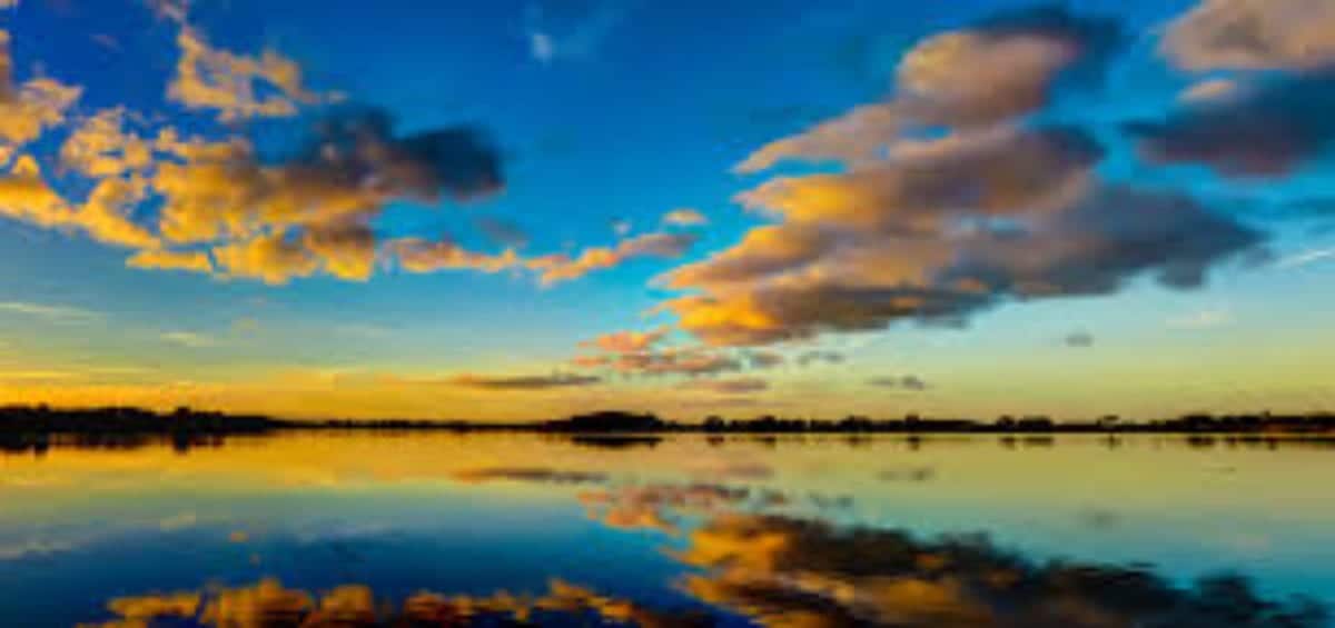 A serene lake at sunset, with vibrant clouds reflecting on the calm water and a bright blue sky transitioning to warm hues.