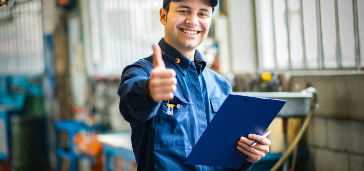 A smiling worker in a blue uniform and cap gives a thumbs up while holding a clipboard in a workshop setting.