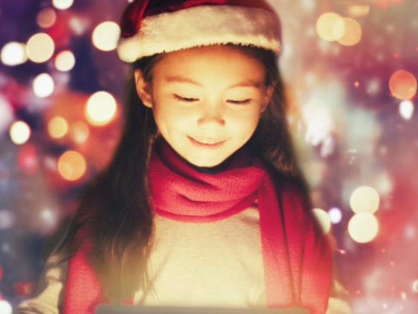 A young girl wearing a Santa hat and red scarf smiles while looking down, with festive lights in the background.