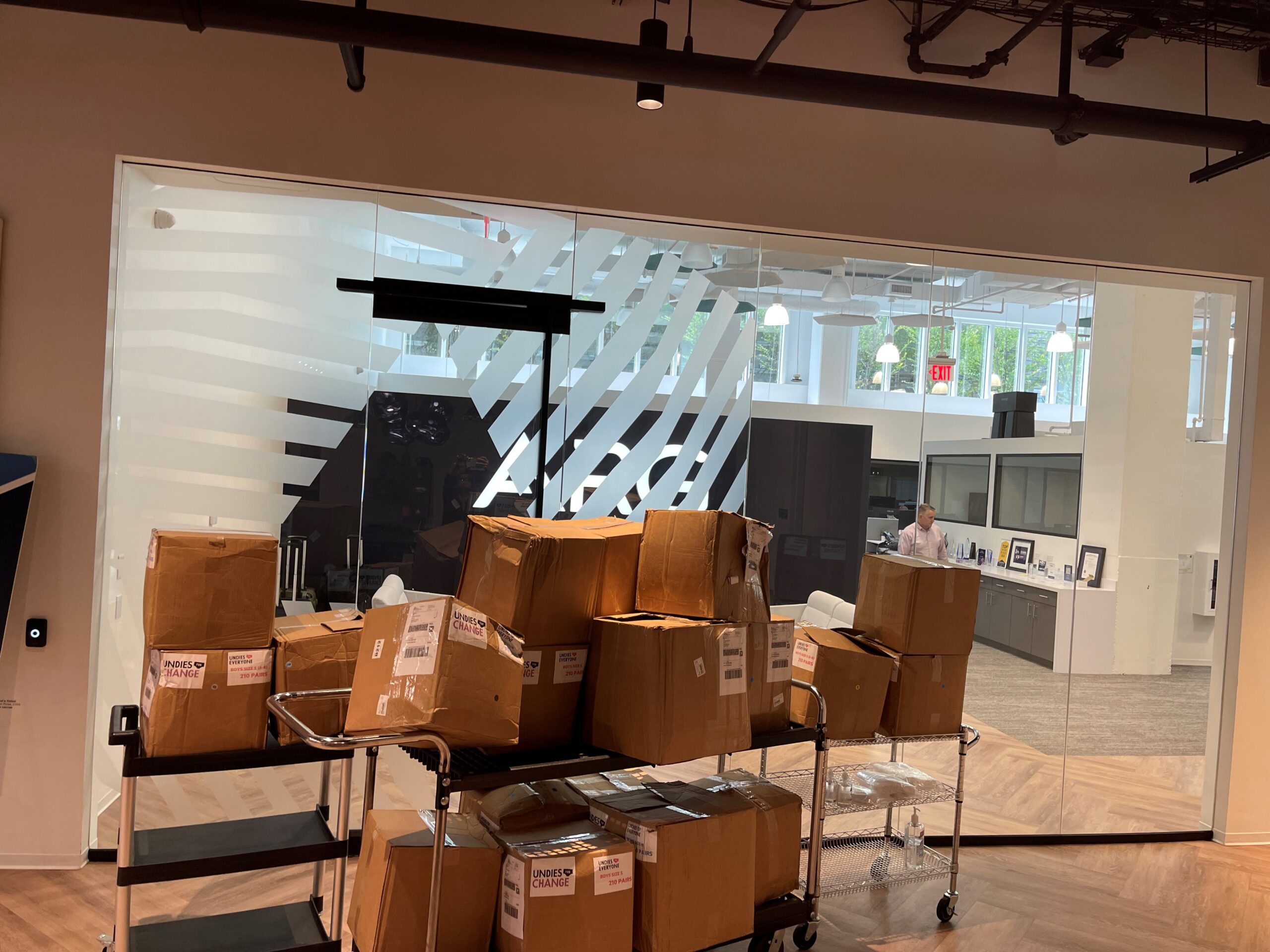 A pile of brown cardboard boxes stacked on a cart is situated in front of a glass-walled office space. A person is visible working at a desk inside the office.
