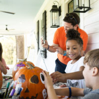 Children and an adult decorating pumpkins with markers and stickers on a porch.