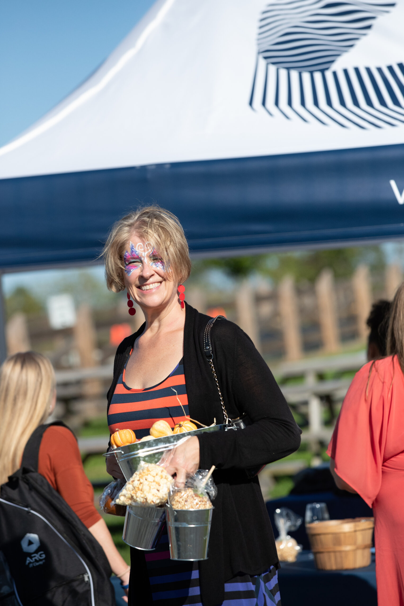 A woman with face paint holds small buckets of popcorn and mini pumpkins at an outdoor event under a tent.