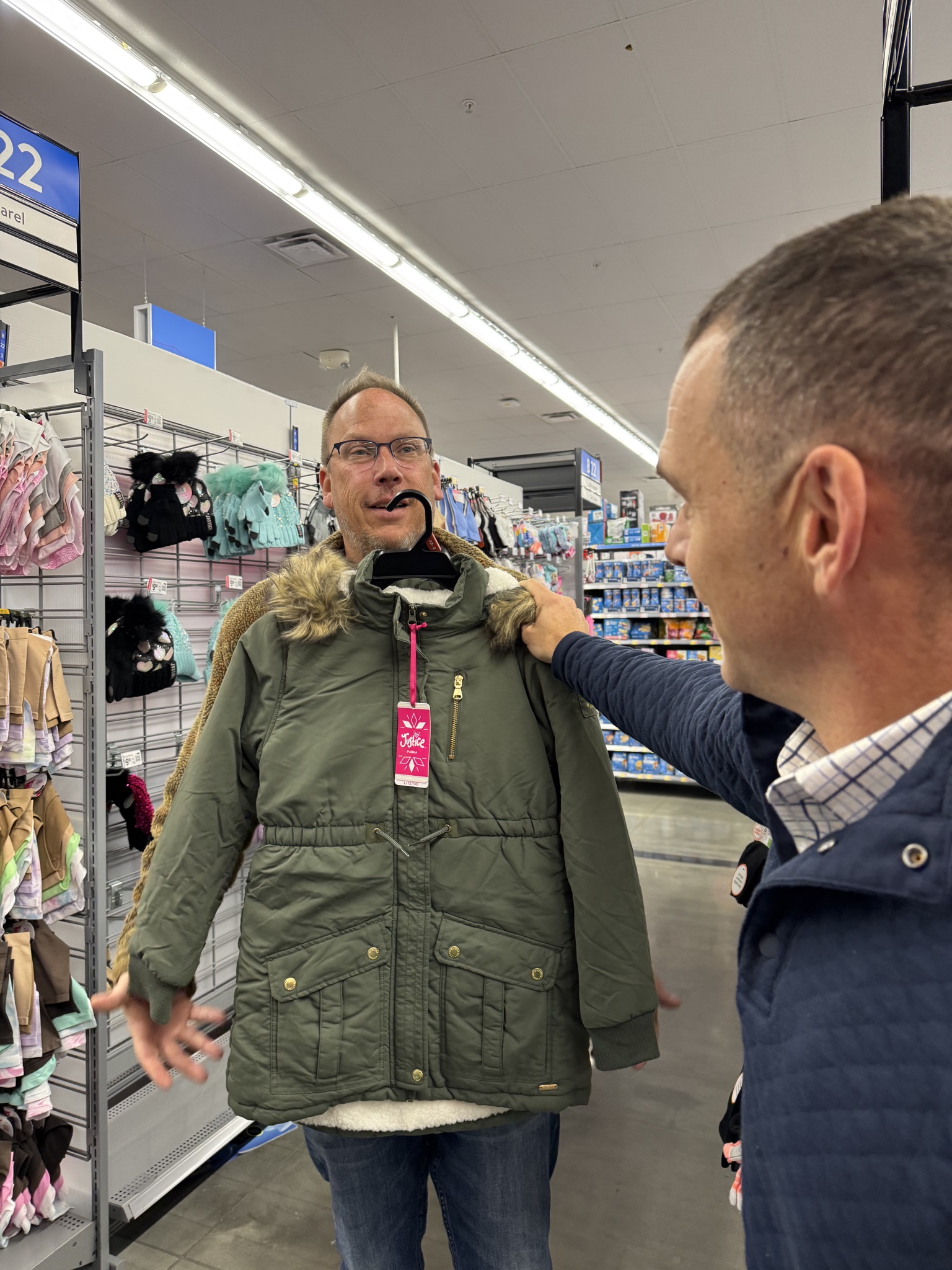 Two people are in a store. One person holds a green coat against the other, who is smiling. They are in a clothing aisle.
