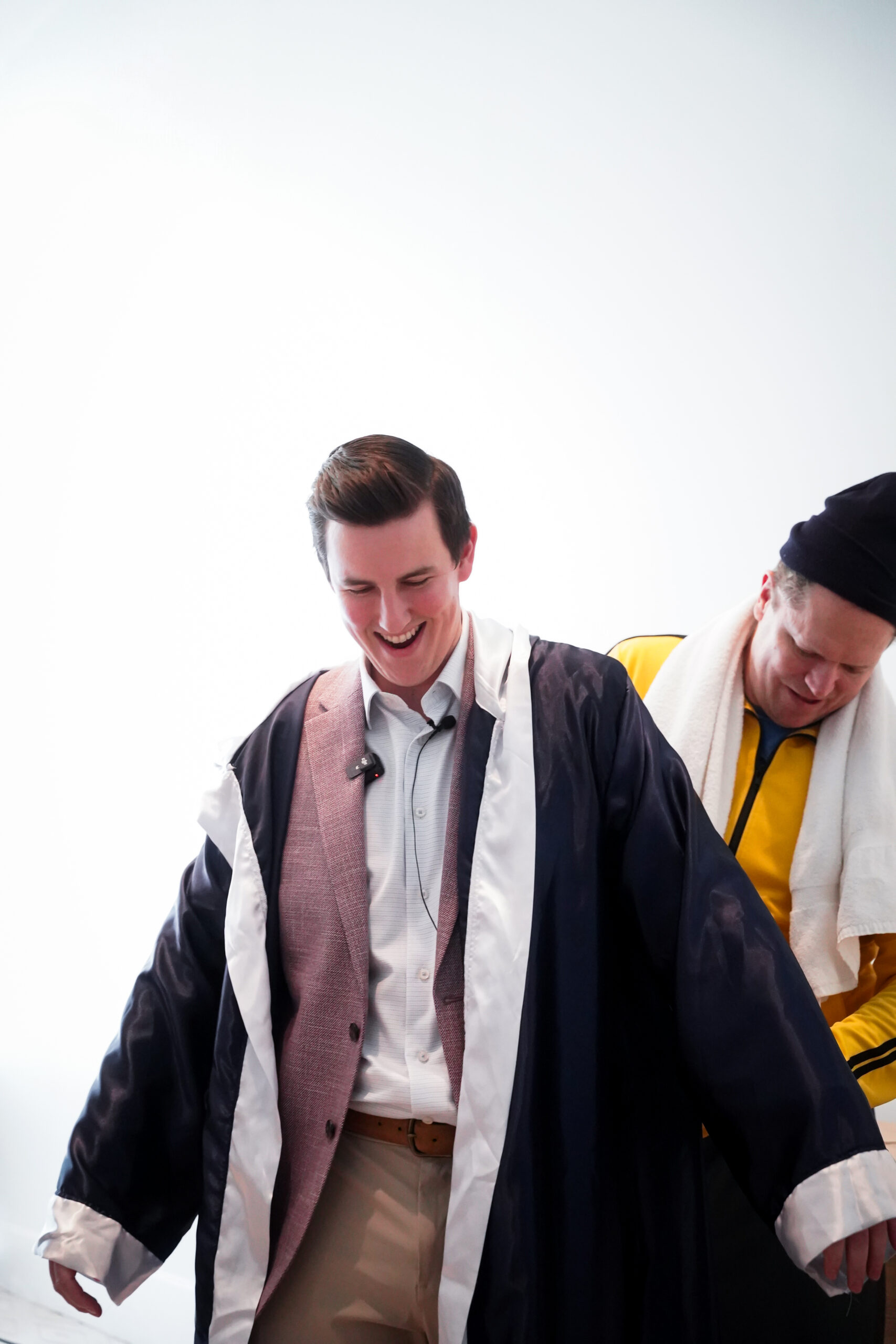 Person smiling while wearing a large black and white coat over a suit, assisted by another person in a yellow shirt and black hat.