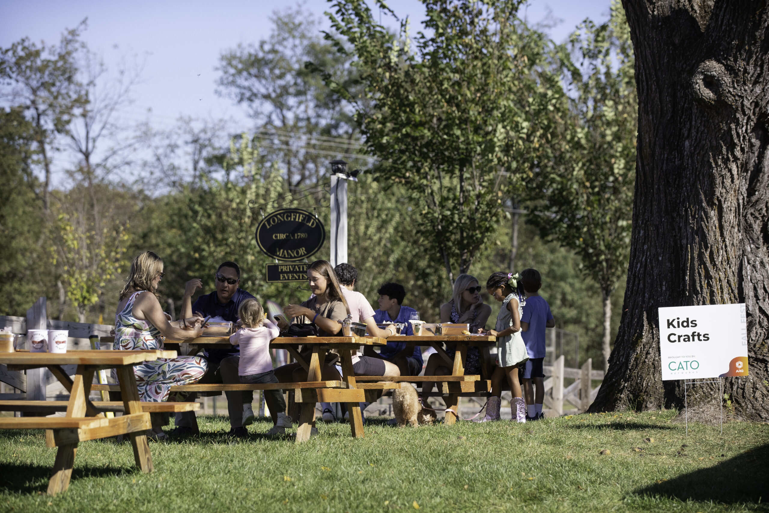 0017_ARG_Fall25 Adults and children sit at picnic tables outside near a tree, with a "Kids Crafts" sign visible on the grass.