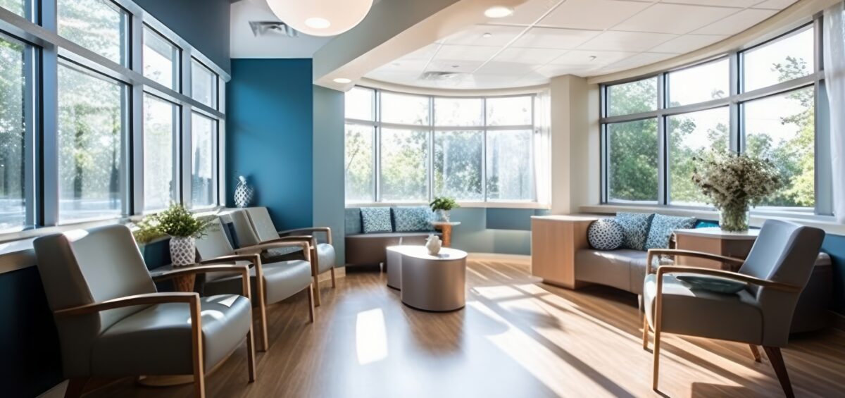 A modern waiting room with large windows, natural light, wooden floors, armchairs, a round table, and plants.